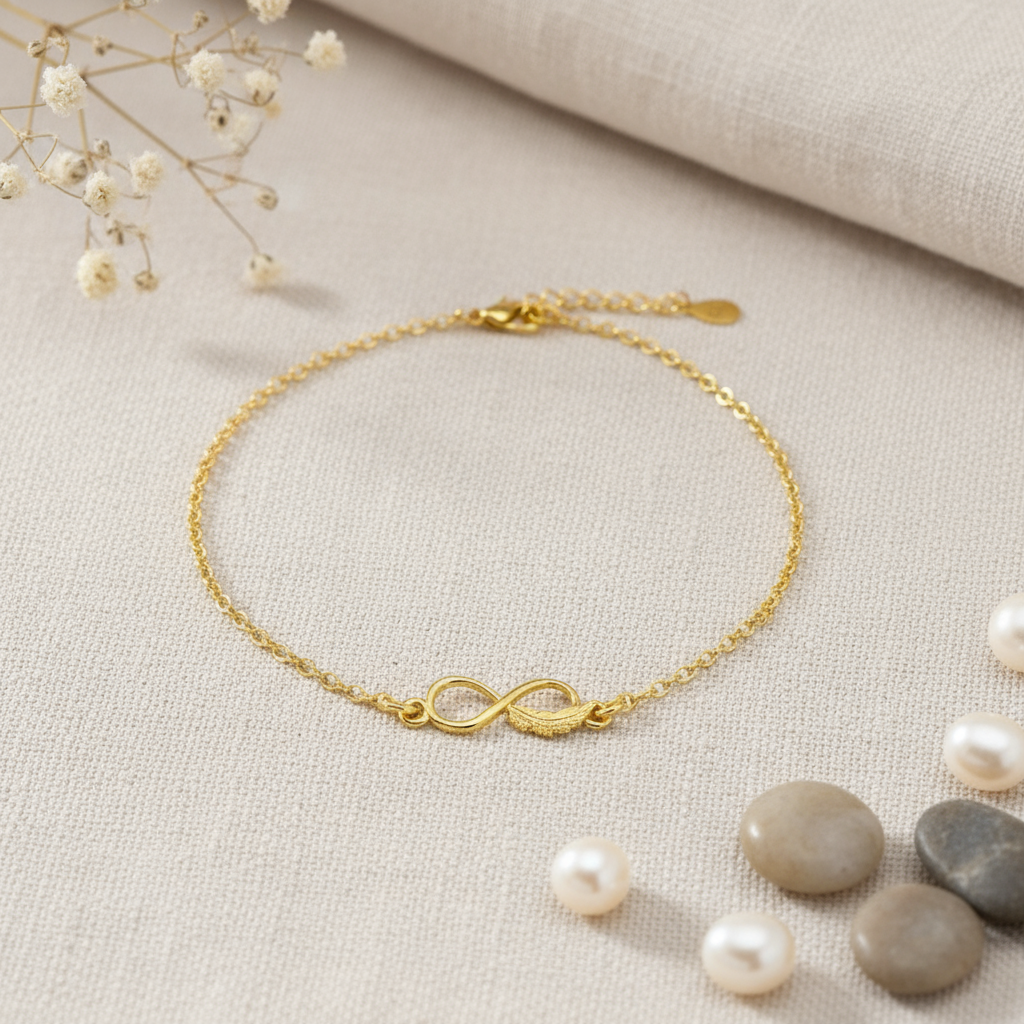Gold ankle bracelet with infinity symbol and feather on a beige fabric background with pearls and stones.
