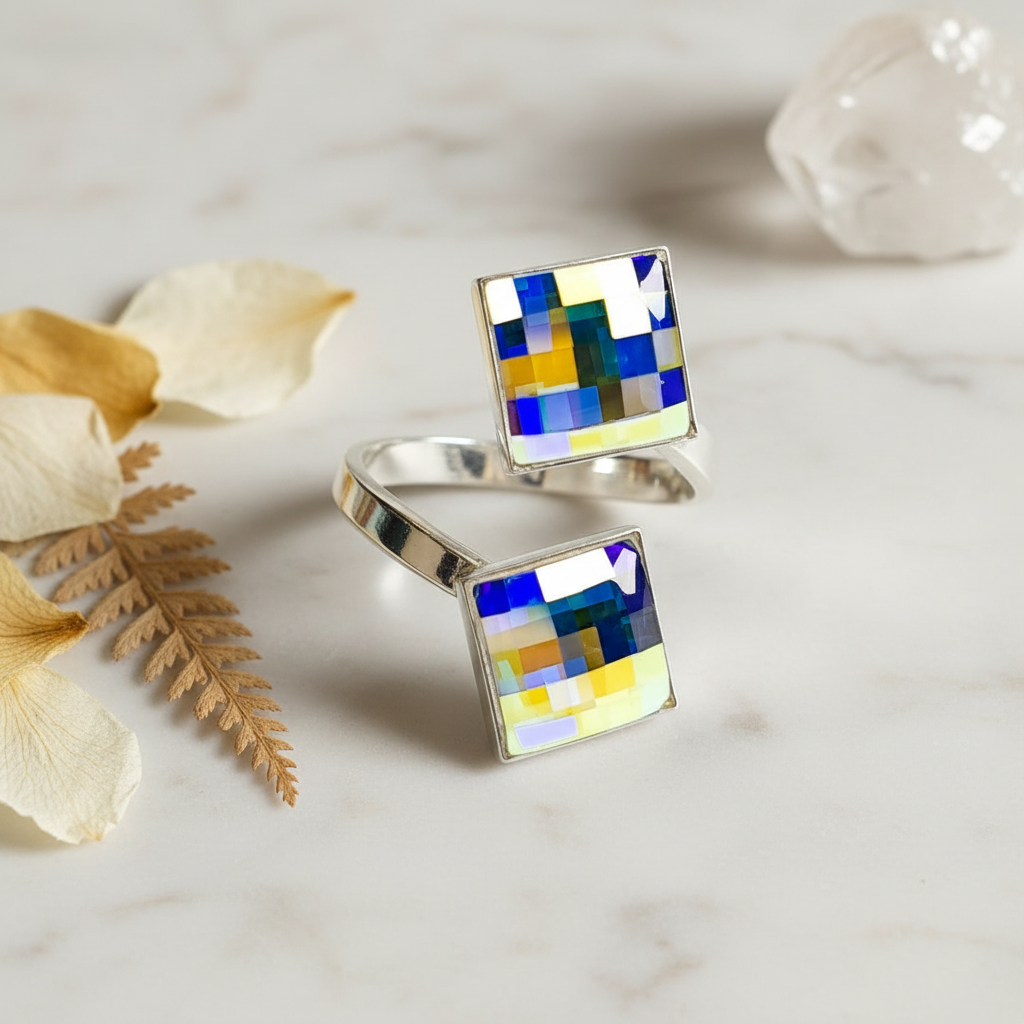 A silver wrap ring with two square, chessboard-patterned aurora borealis-finished crystals on a wooden surface with a green leaf in the background.