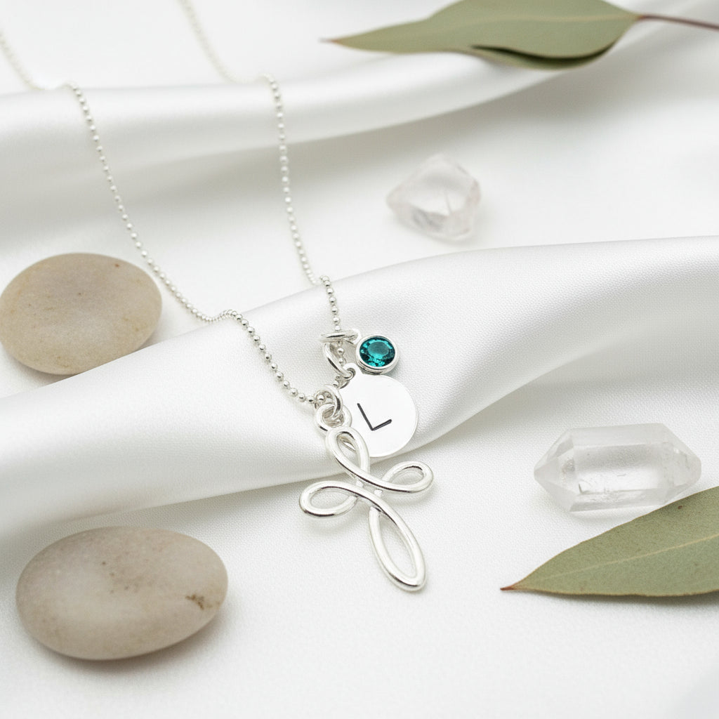 A personalized silver necklace featuring an infinity cross pendant, a disc charm with an engraved initial, and a birthstone charm, displayed on a white background.