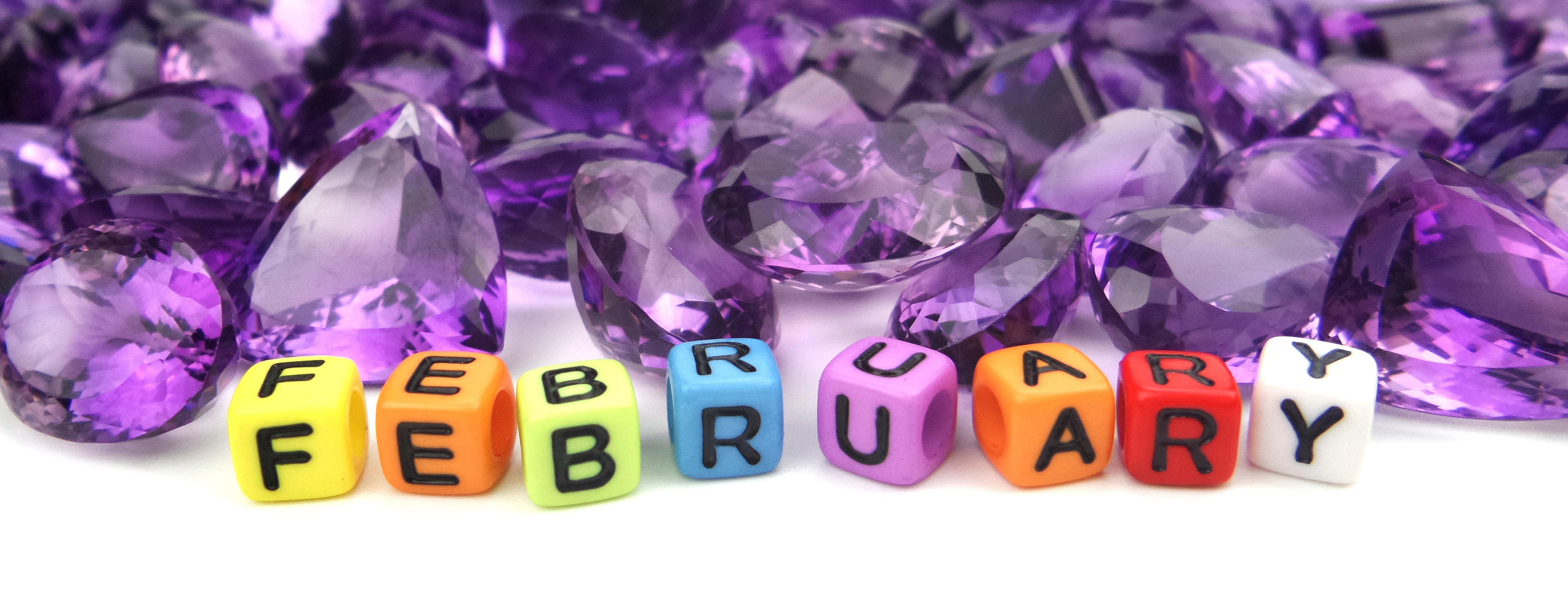 An artistic display of various shaped and sized amethyst crystals on a white background, with the word "FEBRUARY" spelled in colorful bead letters.
