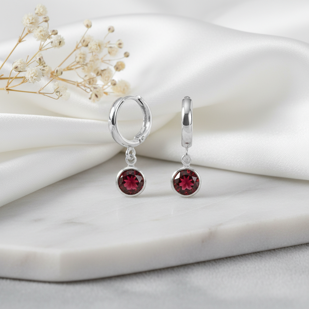 Sterling silver hoop earrings with deep red garnet birthstone charms on a marble surface with dried flowers.