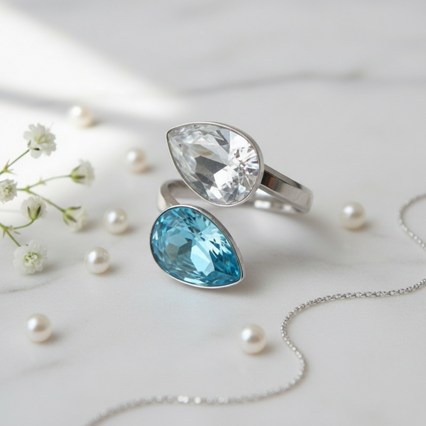 Silver ring with teardrop-shaped blue gemstone on a light background with pearls and flowers.