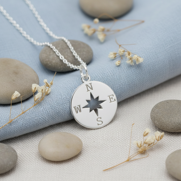 Silver compass necklace on pebbles and dried flowers with a blue fabric background, a gift for your daughter graduation.
