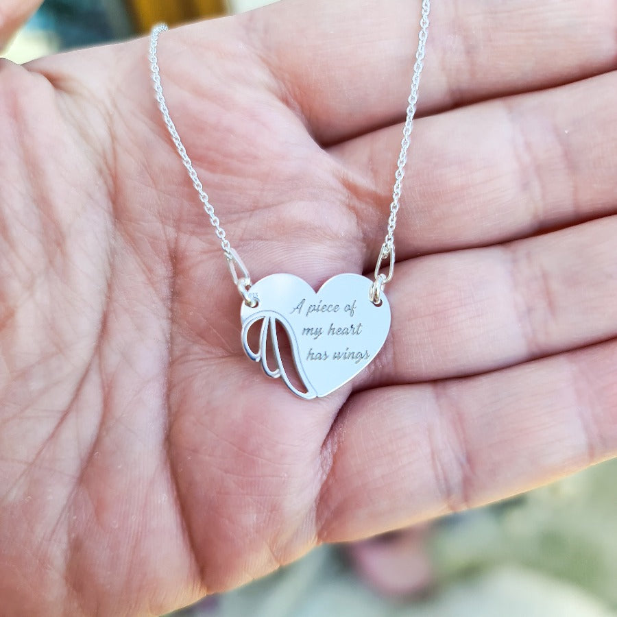 Hand holding a sterling silver Winged Heart Necklace by Magpie Gems, displaying the angel wing pendant and engraved message, with a 45cm chain to illustrate scale.
